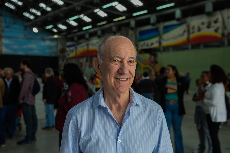 Um homem com um sorriso gentil posa para a câmera, vestindo uma camisa azul clara. Ele está em um ambiente interno com várias pessoas ao fundo, sugerindo um evento social ou uma reunião.