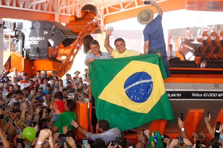 Segurando uma bandeira do Brasil no alto de um trator, Bolsonaro, vestido de polo amarela, acena para o público que está abaixo, ao lado de Tarcísio. Atrás deles, outro trator e um homem de chapéu de palha aparece de costas fazendo uma foto com o celular 