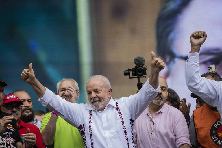 Lula no centro está com braços para cima, fazendo o sinal de positivo com os dedos polegares, olhando para o público,  cercado por outras pessoas, uma aplaude, outra está com os braços para cima 