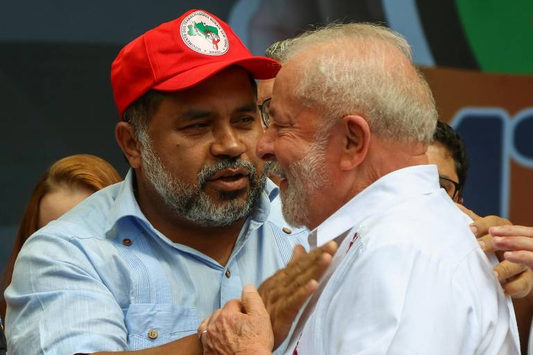 Lula, um homem branco de cabelos ralos e barba da mesma cor, recebe um cumprimento no peito de outro homem, negro de pele clara, barba grisalha e boné vermelho do MST
