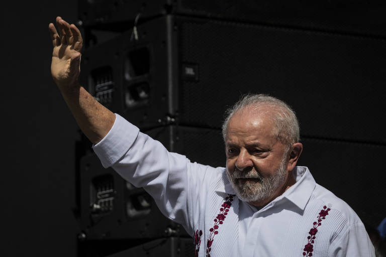 O presidente Lula, de camisa branca, à frente de um fundo preto, ergue a mão direita