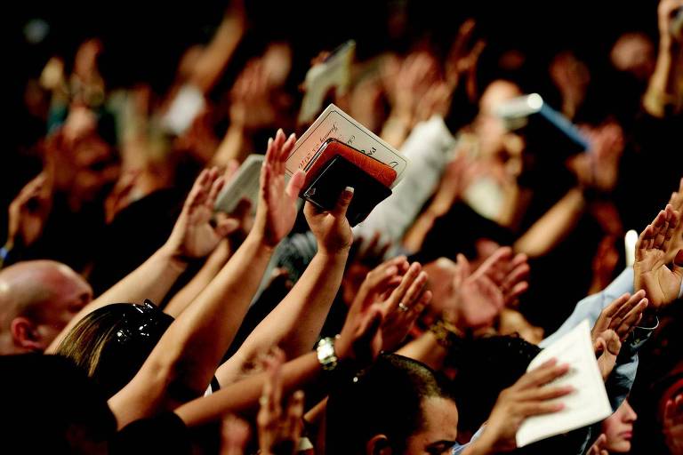 Fiéis evangélicos oram em um culto com os braços erguidos, alguns segurando bíblias