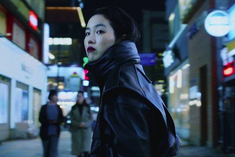 Park, uma mulher jovem, veste casaco preto com a gola alta prendendo seus cabelos. Ela tem as mãos nos bolsos e olha para trás, em uma rua iluminada em Seul