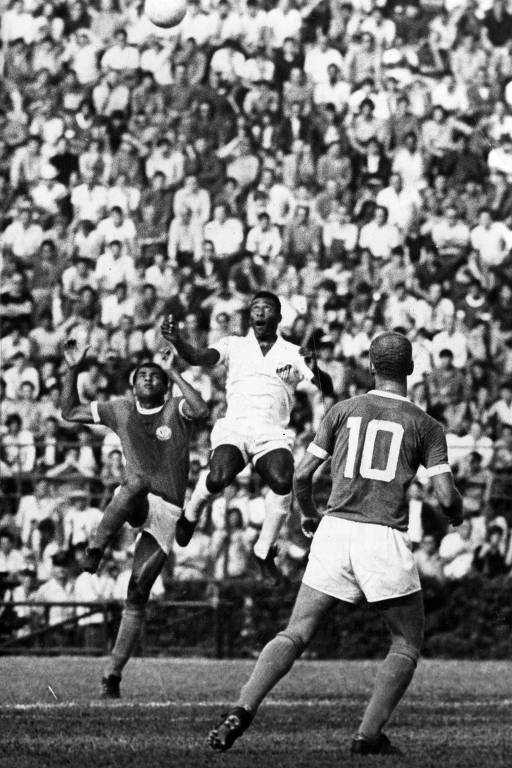 Fotografia antiga, preto e branca, mostra partida de futebol; em campo, no centro, Pelé aparece no ar entre dois jogadores adversários, olhando para a bola acima; ao fundo, uma arquibancada lotada