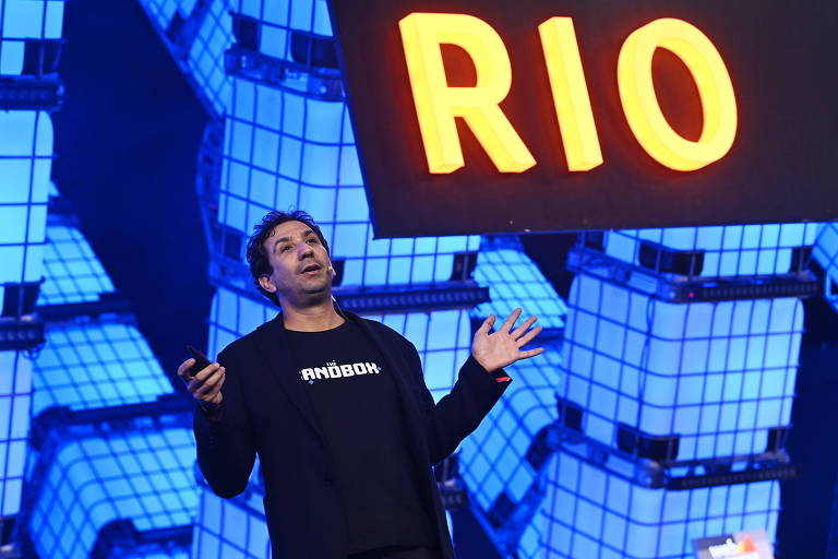 À frente de painel onde se lê a palavra "Rio" iluminada, Arthur Madrid fala gesticulando com os braços abertos. Ele é visto em um ângulo de baixo para cima. Usa uma camiseta com a logo da empresa The Sandbox e uma jaqueta por cima. Ele está com microfone preso à cabeça e segura um passador de slides com uma das mãos. Não tem barba, tem cabeços curtos escuros.