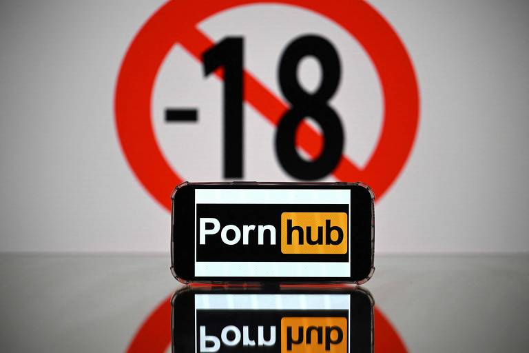 Imagem mostra celular com logo do Pornhub, que é preto e laranja. Atrás, há uma placa com sinal de proibido para menores de 18 anos.