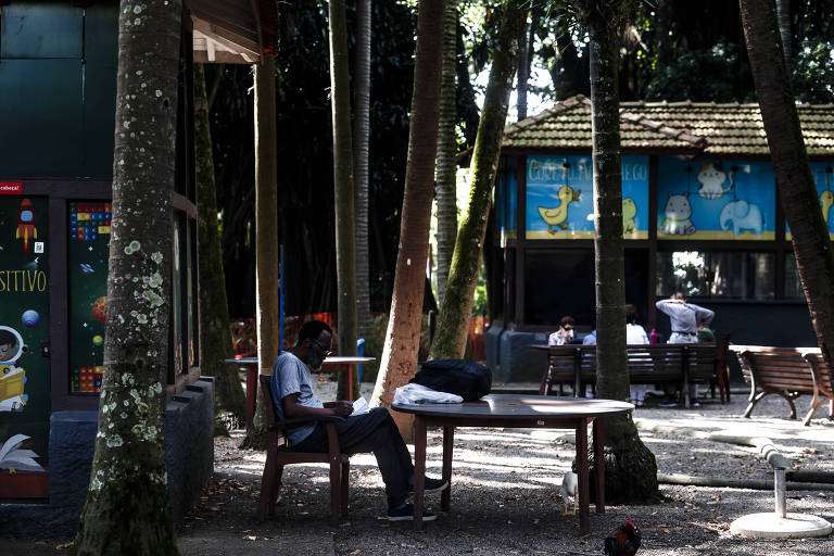 foto de uma pessoa lendo sentada numa cadeira com uma bolsa na mesa e ao fundo uma cabana de leitura com outras pessoas sentadas em volta de uma mesa
