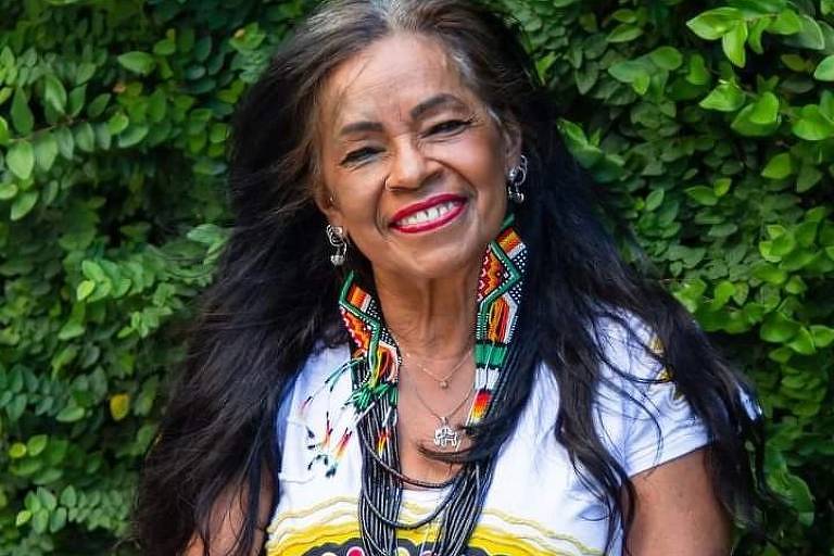 a foto mostra uma mulher indígena de cabelos longos pretos, com alguns fios grisalhos, sorrindo. ela usa um vestido branco com detalhes coloridos, longo brinco com miçangas e colar