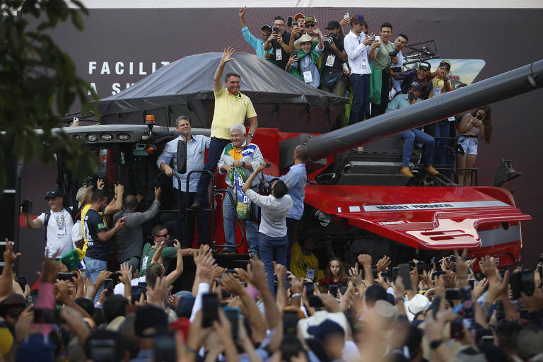 Público, em primeiro plano, está virado para Jair Bolsonaro, que está de camiseta amarela, sobre um trator; ao lado dele está Marcos Pontes e Tarcísio de Freitas