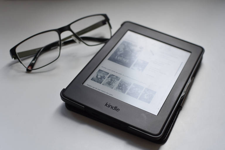 Dispositivo Kindle, preto, com tela em preto e branco, ao lado de óculos.