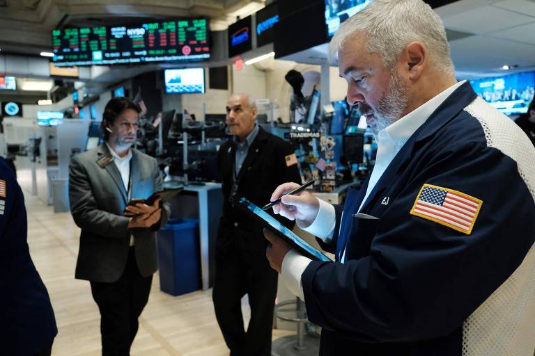 Investidores trabalham no térreo da Bolsa de Nova York, nos Estados Unidos. Na imagem estão três homens: o da direita, veste um casaco, com a bandeira dos Estados Unidos, na manga, e é grisalho; o do meio é calvo e veste jaqueta de moletom preta sobre camisa azul; o da esquerda tem cabelos castanhos e veste terno.