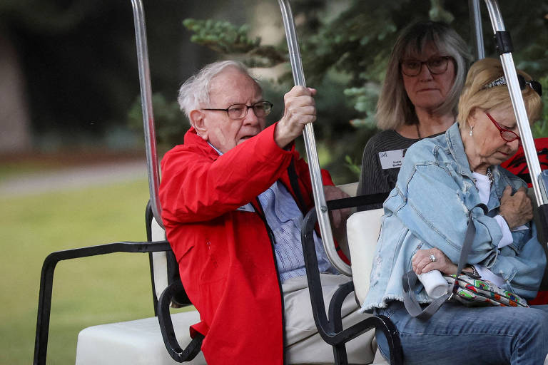 O CEO da Berkshire Hathaway, Warren Buffett, participou do Congresso de Mídia de Sun Valley, em Idaho, Estados Unidos. Buffett, 92, é um homem de cabelos brancos e ralos, vestido em casaco vermelho e camisa branca. Ele está em um carrinho de golfe, ao lado de duas senhoras grisalhas.