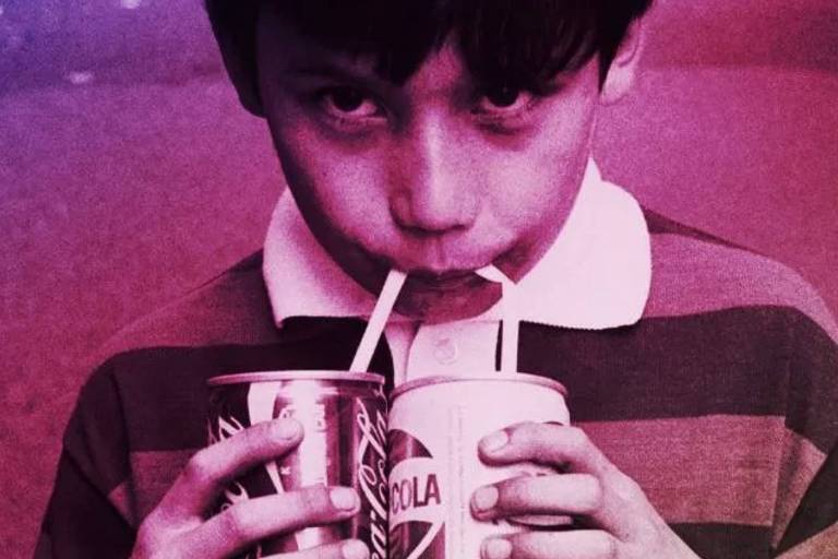 Foto colorida em tons magenta mostra menino tomando refrigerante em duas latas, com canudos