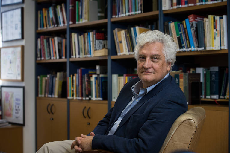 Homem branco, de cabelo branco, sentado em frente a uma estante cheia de livros