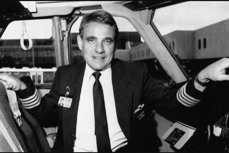Em foto preta e branco, homem com uniforme de piloto tira foto em uma cabine de avião