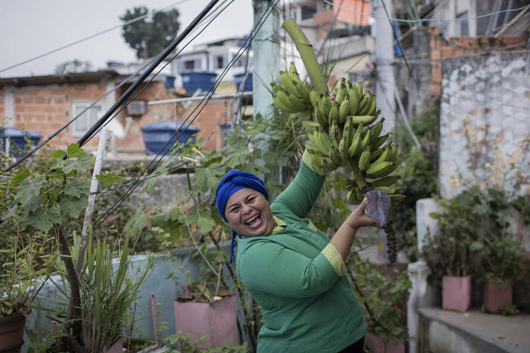 Imagem colorida mostra a chef de cozinha Regina Tchelly apontando para a horta da Favela Orgânica, no morro da Babilônia, na zona sul do Rio de Janeiro. Ela sorri, veste uma roupa verde com detalhes em amarelo e um turbante azul na cabeça.