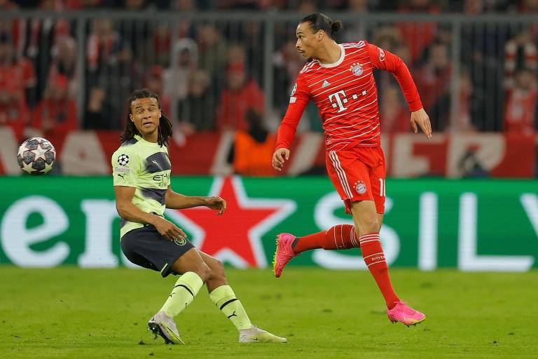 Ake, do Manchester City, e Sané, do Bayern, disputam bola em confronto da Champions; ao fundo placa da Heineken Silver