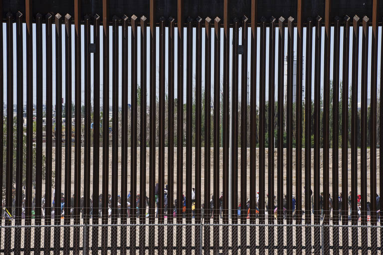 Atrás de uma cerca de fronteira, imigrantes esperam para passar pela segurança dos EUA