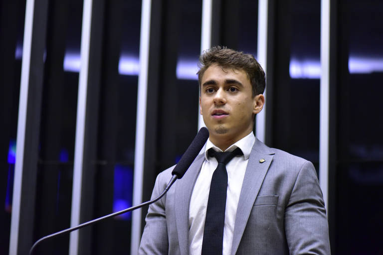 Nikolas, um rapaz jovem, branco, cabelos castanhos e vestido de terno cinza com gravata azul escuro e camisa branca. Ele está de pé falando ao microfone de mesa