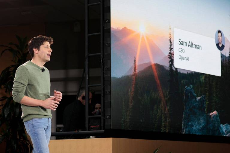 Chefe-executivo da OpenAI, Sam Altman, durante evento de anúncio da integração do ChatGPT ao buscador da Microsoft Bing. Altman é um homem branco de suéter verde a calças jeans. Ele está à esquerda de um telão.