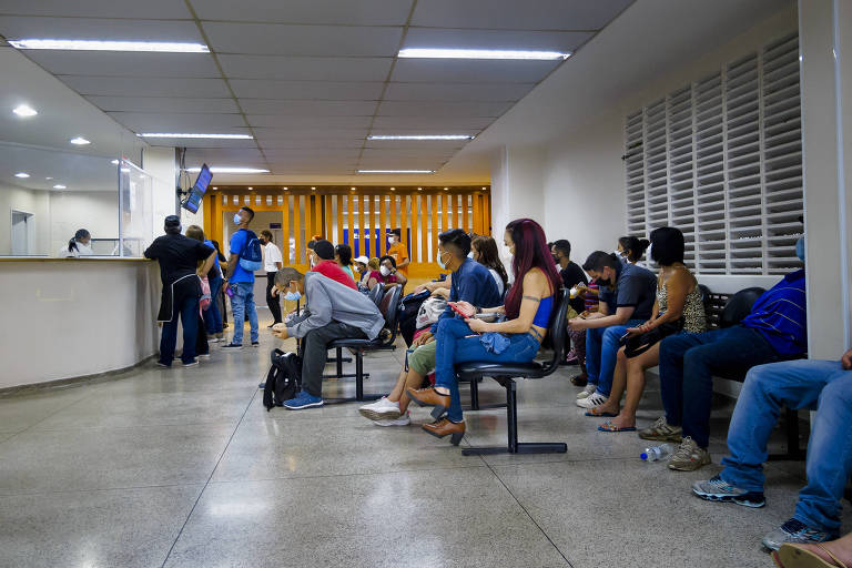 Pacientes aguardam sentados em cadeiras de saguão, onde há um balcão de atendimento e uma tela com o número de chamada