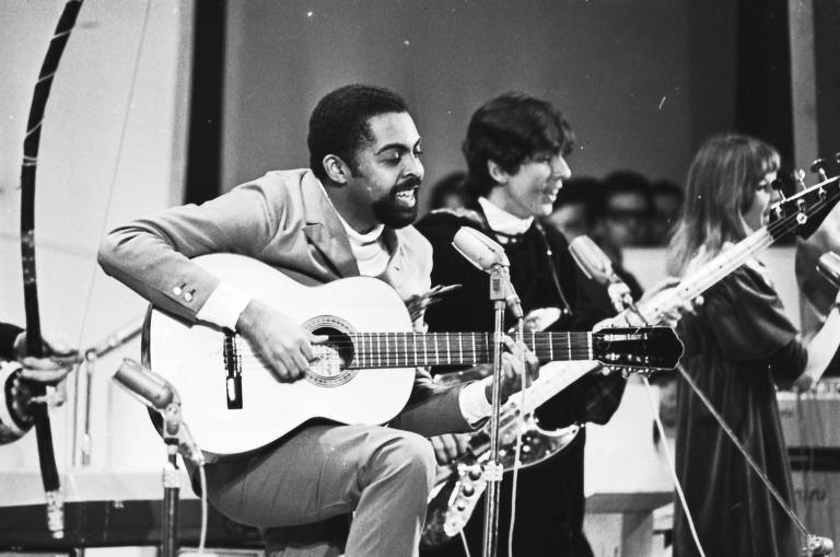 fotografia em preto e branco mostra Gilberto Gil sentado, tocando violão e cantando ao microfone 