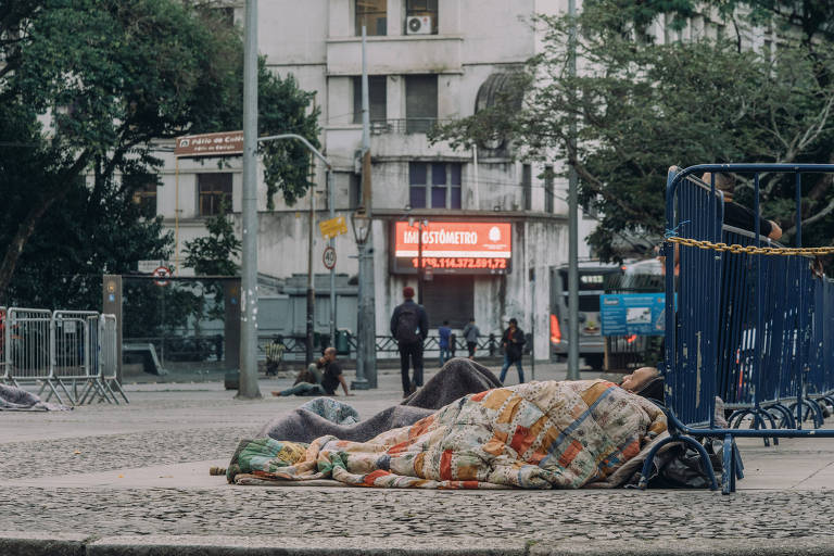 foto mostra pessoas dormindo em praça com o corpo inteiro coberto. ao fundo, gradis azuis do lado direito, e pessoas circulando mais ao fundo