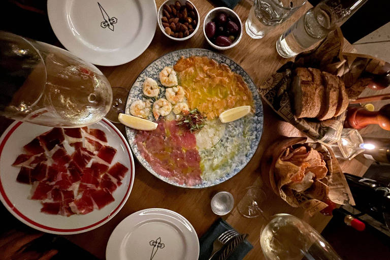 A foto mostra uma mesa vista de cima com pratos de petiscos, como presunto espanhol, azeitonas, castanhas, torradas e um prato de pescados e camarões ao centro