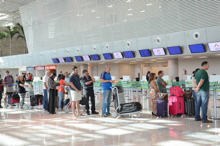 Fila de passageiros no espaço de check-in do aeroporto de São Gonçalo do Amarante (RN), eles usam camisas de cor preta, azul, verde, as malas são vermelhas, azuis e pretas e o local é bem iluminado e tem o piso claro