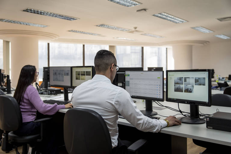 Imagem colorida mostra uma mulher, à esquerda, e um homem, sentados de costas e trabalhando em computadores