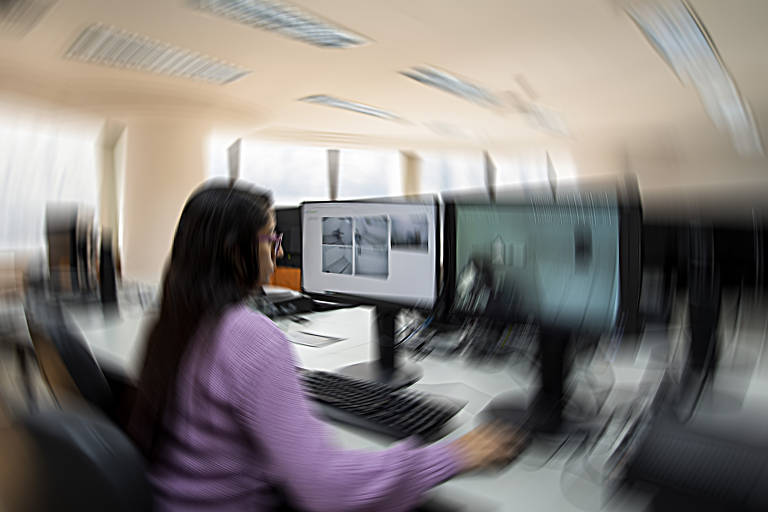 Imagem colorida mostra uma mulher sentada de costas e trabalhando em computadores