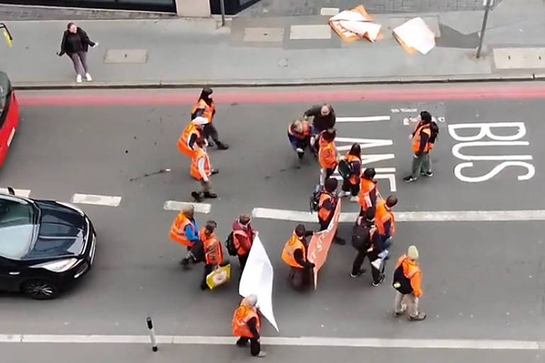 foto mostra grupo de manifestantes com roupas laranjas caminhando no meio da rua. eles olham para um homem de casaco preto que empurra uma das ativistas para o chão