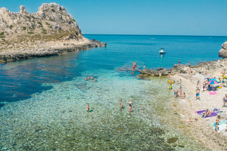 Praia Cala Camarro, na ilha Levanzo, no meio do mar Mediterrâneo. À direita, há uma faixa de areia com banhistas e turistas descansando, e algumas pessoas na porção mais rasa do mar. Um pouco distante da costa, há uma pequena ilha rochosa