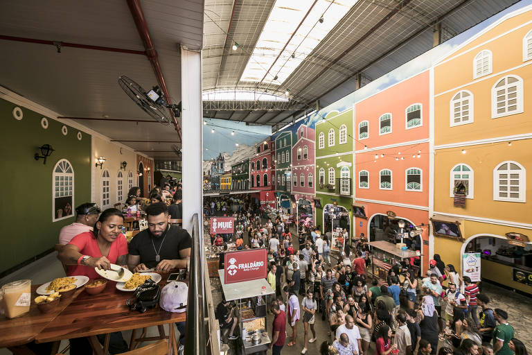 A imagem mostra uma feira de comida em um ambiente vibrante, com barracas e edifícios coloridos ao fundo. Há um grande número de pessoas caminhando e interagindo no espaço, enquanto algumas pessoas estão sentadas em uma mesa à esquerda, com pratos de comida. O teto é de vidro, permitindo a entrada de luz natural, e as paredes dos edifícios são pintadas em cores vivas como verde e laranja.