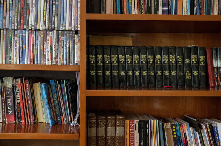 Em fotografia frontal, estante de madeira com bíblias, livros e DVD.