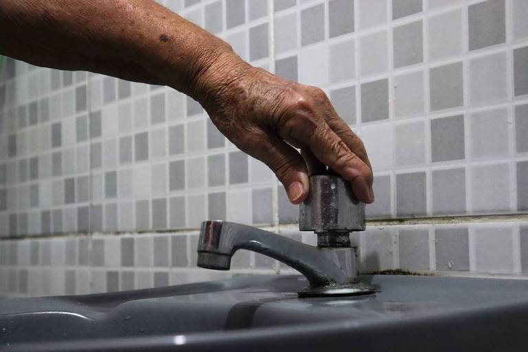 A foto mostra uma torceira sendo acionada pela mão de uma pessoa idosa e de pele negra; a água não é entregue; o ambiente é de um banheiro
