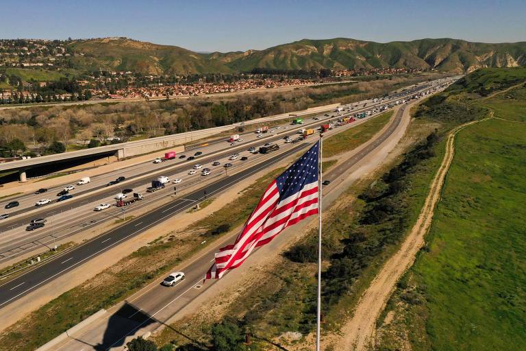 Fluxo de veículos em estrada na Califórnia, nos EUA, com bandeira do país hasteada em mastro na lateral
