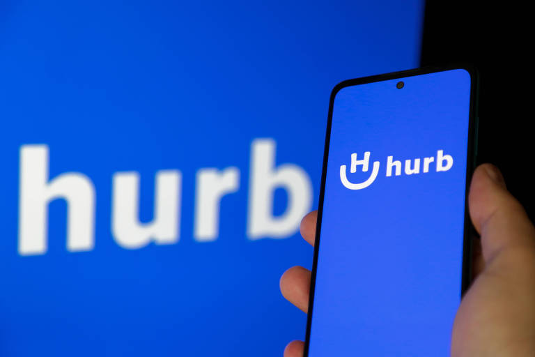 Mão segura smartphone com logo do Hurb, à frente de banner com logo da plataforma.