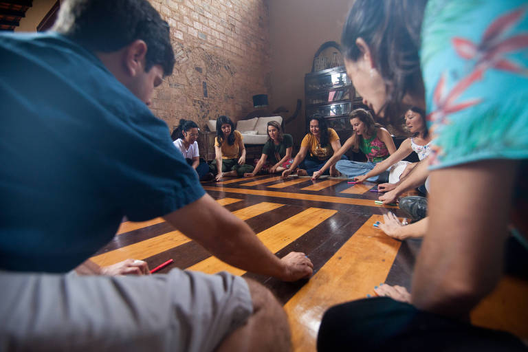 Grupo de pessoas está sentado no chão em forma de círculo e participam de uma dinâmica