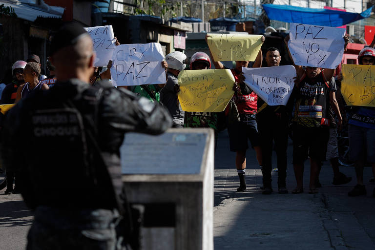 pessoas exibem cartazes de protesto pedindo paz. em primeiro plano, desfocado, um policial de costas