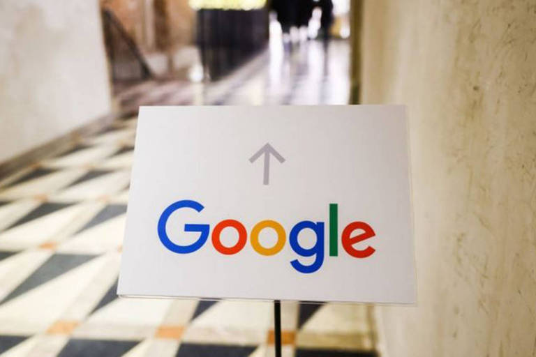 Imagem mostra placa com logo do  Google abaixo de uma seta que indica: "siga adiante." Paredes são beges e pisos  de granito, branco e preto