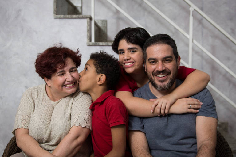 Família posa para foto sentada em sofá; o menino beija a mãe; a filha mais velha abraça o pai