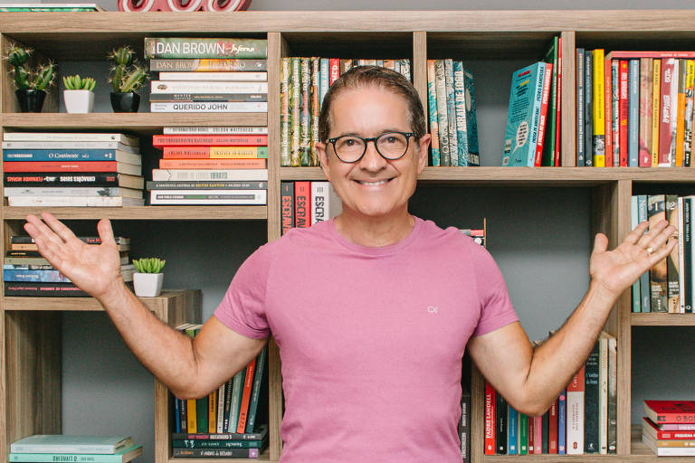 Jornalista Carlos Tramontina, de camiseta rosa, sorri e abre os braços olhando para a câmera; ao fundo, uma estante cheia de livros e um luminoso onde se lê a palavra "Joy" (Alegria, em inglês)