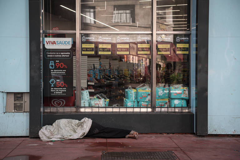 Fotografia colorida de pessoa negra dormindo em uma calçada, diante da vitrine de uma loja, com a cabeça coberta