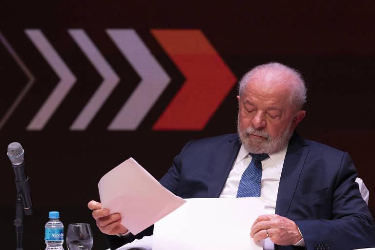 O presidente Luiz Inácio Lula da Silva (PT) compareceu ao fórum Indústria Forte, País Forte, da Fiesp. Lula veste paletó marinho, camisa branca e gravata azul. Ele segura papéis e está à frente de parede preta com setas brancas e vermelhas