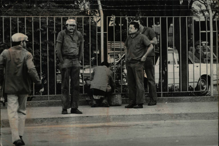 Foto em preto e branco mostra gradil em frente ao qual estão três homens de jaqueta e boné