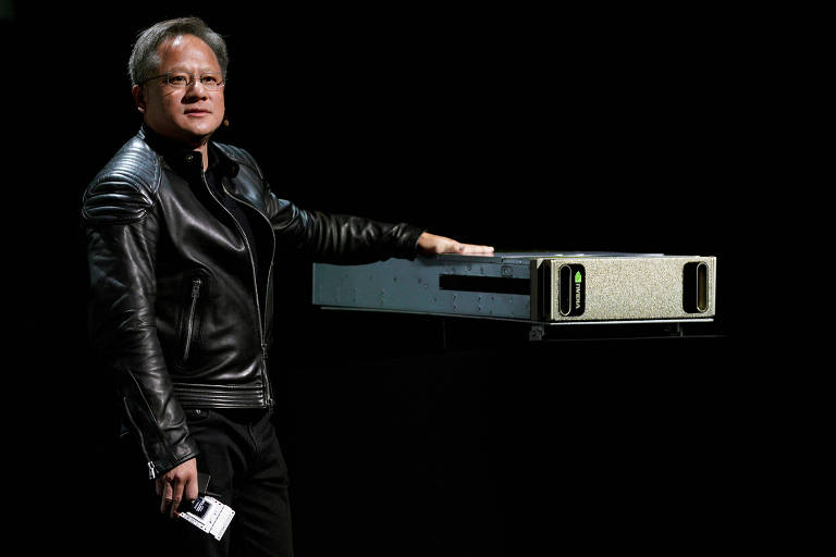 O presidente-executivo da Nvidia, Jensen Huang, mostra o supercomputador de inteligência artificial DGX AI, de sua empresa, em evento de tecnologia realizado em Las Vegas, EUA. Huang veste camisas pretas, calças pretas e jaqueta de couro escura. Ele está ao lado do gabinete do computador, com a mão sobre o tampo.