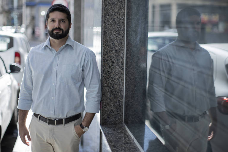 Imagem colorida mostra Virgilio Ferreira, um homem branco de 35 anos. Ele veste uma camisa social azul claro e uma calça bege. Usa barba e tem a imagem refletida em uma porta de vidro à direita.