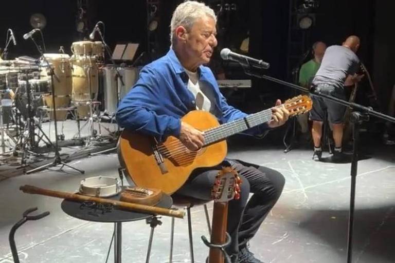 Chico Buarque, sentado no banquinho, canta e toca violão; ele usa calça e camisa jeans