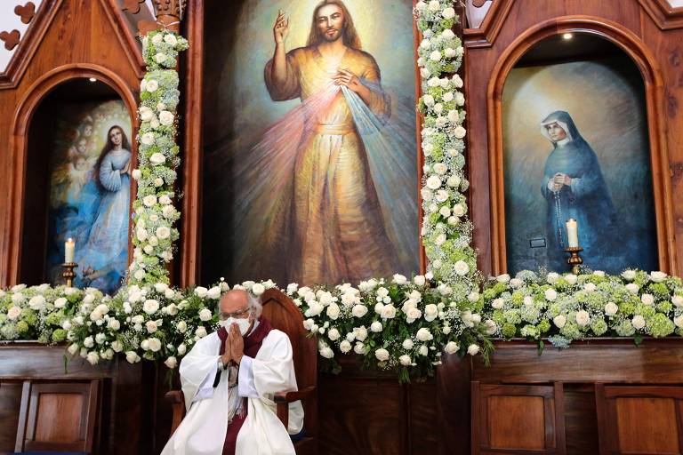 Um cardeal, vestido de branco e sentado, celebra missa em uma igreja com uma imagem de Jesus acima de si